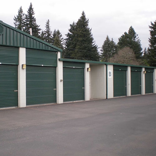 a series of metal roll up self storage unit doors with paved parking in front of the drive-up units and evergreen trees in the background