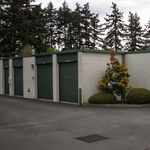 a row of self storage units with green metal roll up doors and paved parking in front of the units with evergreen trees in the background